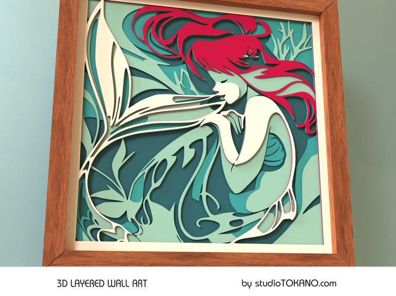 Op de afbeelding: Een 3D gelaagd wandkunstwerk met een zeemeermin met golvend rood haar, een witte staart en een turquoise en groene achtergrond. De kunst is ingelijst in een natuurlijk houten frame.