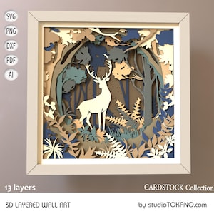 May include: A framed 3D layered wall art piece featuring a white deer in a forest scene. The artwork uses multiple layers of paper in shades of blue, green, and tan to create depth. The frame is a light beige color. The text "13 layers" and "3D LAYERED WALL ART" are visible.