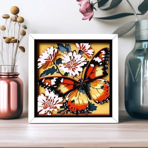 Peut inclure: Un cadre carré blanc contenant une œuvre d'art en papier multicouche représentant un grand papillon orange, noir et jaune au centre, entouré de fleurs blanches et de feuilles bleu foncé sur un fond jaune uni, créant un effet de relief.