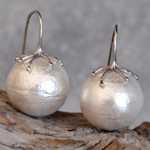 May include: A pair of silver-toned drop earrings. Each earring features a large, round, off-white pearl-like bead suspended from a decorative silver element. The silver element has a branch-like design with small, round accents.