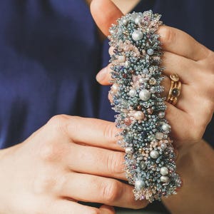 Puede incluir: Una pulsera tipo brazalete ancha y texturizada, densamente adornada con una mezcla de pequeñas cuentas, cristales y perlas en tonos azul polvoriento, rosa suave, plata y gris iridiscente, creando una textura superficial muy detallada.