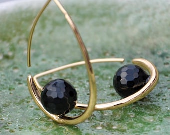 May include: A pair of gold-colored hoop earrings, each featuring a faceted black bead. The earrings have a modern, minimalist design, with a curved shape that cradles the black beads. The earrings are set against a green background.