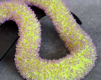 May include: A beaded accessory in a loop shape, featuring a vibrant yellow base color with a scattering of small, pink beads. The texture appears fuzzy and three-dimensional, creating a playful and eye-catching design. The accessory is set against a neutral gray background.