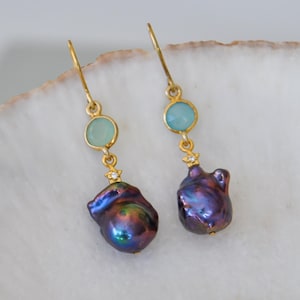 May include: A pair of gold-toned drop earrings. Each earring features a light blue gemstone, a small star-shaped accent, and a large, iridescent, dark purple baroque pearl. The earrings are displayed on a white surface.