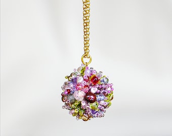 May include: A gold-colored chain necklace with a round pendant. The pendant is a cluster of colorful beads in shades of pink, purple, green, and red. The beads are of various sizes and shapes, creating a textured, eye-catching design.