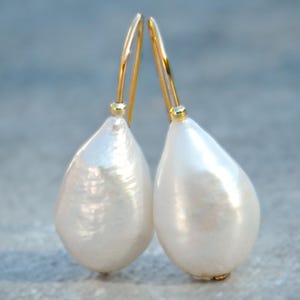 Op de afbeelding: Een paar druppelvormige oorbellen met grote, peervormige witte parels. De oorbellen hebben goudkleurige, gebogen oorhaken. De parels hebben een glanzend, glad oppervlak dat licht reflecteert.