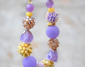 May include: A handcrafted necklace featuring a variety of beads in shades of purple, yellow, and gold. The necklace includes round, faceted, and textured beads, as well as flower-shaped pendants. The design is intricate and colorful, suitable for a special occasion.