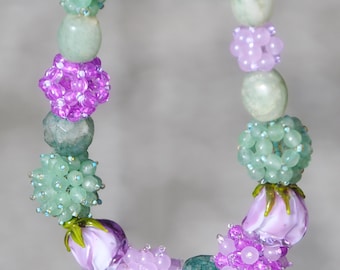 May include: A handcrafted necklace featuring a variety of beads in shades of green and purple. The necklace includes oval-shaped green beads, clusters of small green beads, and spherical purple beaded accents, along with glass flower beads.