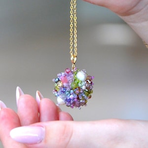 Könnte beinhalten: Eine goldfarbene Halskette mit einem Anhänger aus bunten Perlen. Der Anhänger hat eine kugelförmige Form mit einer Mischung aus rosa, lila, grünen und weißen Perlen. Die Halskette wird von einer Hand mit lackierten Nägeln gehalten.