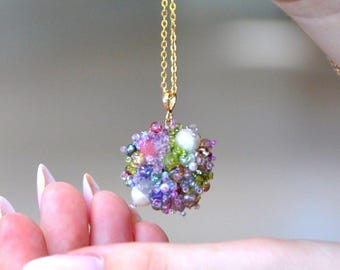 Puede incluir: Un collar de cadena dorada con un colgante esférico compuesto por cuentas de colores. El colgante tiene forma de esfera, con cuentas rosas, moradas, verdes y blancas. El collar lo sostiene una mano con esmalte de uñas rosa.