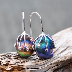 Pode incluir: Um par de brincos de pérolas iridescentes. Cada brinco apresenta uma pérola em forma de gota com uma mistura vibrante de cores, incluindo azul, verde e dourado. As pérolas são suspensas por ganchos prateados com um design único e estilizado.