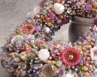 May include: A handcrafted, beaded wreath in a crescent shape. The wreath is densely covered with a variety of beads in shades of pink, purple, green, and gold. Large pearls and faceted beads are interspersed throughout the design, creating a textured, decorative piece.