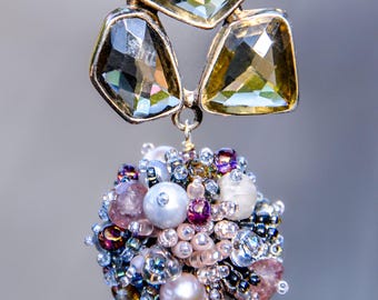 May include: A handcrafted pendant necklace featuring a cluster of colorful beads and pearls. The pendant is adorned with a trio of faceted, geometric-shaped crystals in shades of gold and gray. The necklace is suspended from a delicate chain.