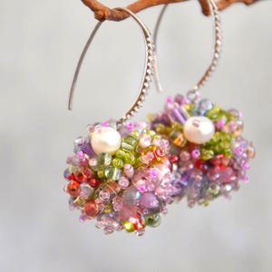 May include: A pair of drop earrings featuring spherical clusters of multi-coloured glass beads in shades of pink, green, and purple. Each earring is accented with a single white pearl and hangs from a textured silver-toned metal hook wire.