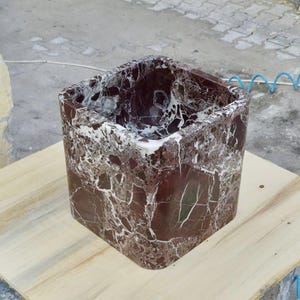 Può includere: Un lavandino quadrato in marmo con angoli arrotondati. Il lavandino è di un colore bordeaux intenso con venature bianche. Il lavandino è appoggiato su una superficie di legno chiaro.
