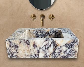 Puede incluir: Un lavabo de baño de mármol blanco con forma rectangular y un patrón único e irregular de vetas grises y doradas. El lavabo está montado en la pared y tiene un grifo con dos manijas.