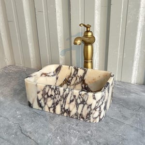 May include: A rectangular marble sink with a white, pink, and purple marbled pattern. The sink is sitting on a grey countertop. A gold faucet is mounted above the sink.