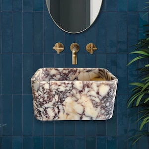 May include: A rectangular, white marble sink with brown and purple veining. The sink is mounted on a blue tiled wall with gold-toned faucets.