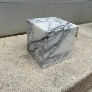 Pode incluir: Um cubo de mármore branco com veios cinzentos. O cubo está sobre uma superfície de concreto cinza.