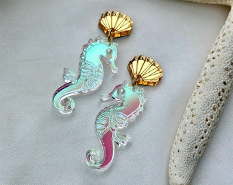 May include: A pair of iridescent seahorse earrings with gold-colored shell-shaped tops. The seahorses have a gradient of colors, including pink, blue, and green. The earrings are displayed on a white surface with a starfish.