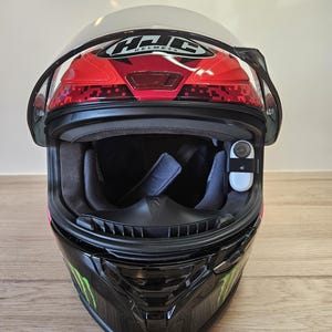 Puede incluir: Un casco de moto negro con un logotipo de Monster Energy verde y negro. El casco tiene una visera transparente y un interior rojo. Una pequeña cámara negra está unida a la parte superior del casco.