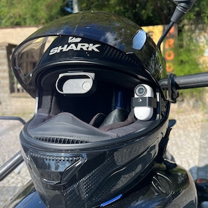 以下が含まれることがあります： 黒いオートバイのヘルメットで、クリアなバイザーと前面に「SHARK」という文字が印刷されています。ヘルメットの側面に小さな白いアクションカメラが取り付けられています。