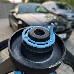 Può includere: Un volante nero con un anello adattatore azzurro e un mozzo centrale nero. Il volante è tenuto davanti a uno sfondo sfocato con auto e alberi.