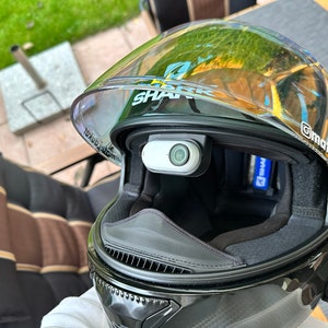 以下が含まれることがあります： クリアバイザーと内蔵カメラ付きの黒いオートバイヘルメット。ヘルメットには側面にSharkのロゴとバイザーにMatejdukátのステッカーがあります。