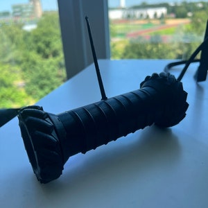 May include: A black 3D printed robot with a long antenna. The robot has a cylindrical body with a textured surface and a round base.