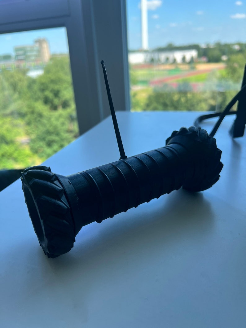 May include: A black 3D printed robot with a long antenna. The robot has a cylindrical body with a textured surface and a round base.