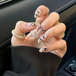 May include: A hand with long, almond-shaped nails. The nails are painted with a nude base and black abstract designs. The hand is adorned with multiple gold rings, including a thick, diamond-studded band. The background is a dark interior.