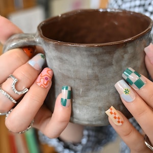 May include: A person holds a grey ceramic mug with a brown interior. Their nails are decorated with various patterns, including green and white checks, orange swirls, and a small yellow flower. Silver rings are worn on the fingers.