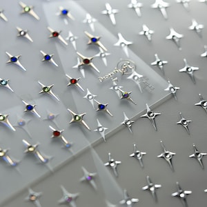 May include: Silver star-shaped nail art decorations with colorful gemstones. The nail art stickers have a four-point star design with a central gem in various colors, including red, blue, green, and clear. The stars are arranged on a clear sheet.