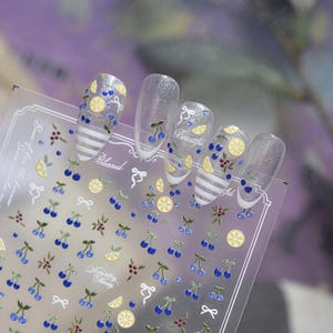 May include: A sheet of adhesive nail art stickers featuring small blue cherries, yellow lemon slices, and white bows. The sheet includes five clear, almond-shaped press-on nails decorated with these motifs, some with white horizontal stripes.
