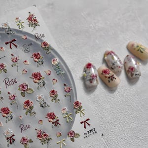 May include: Nail art stickers with rose designs in red, pink, and white, and small bows. The word "Rose" is printed on the sheet. Several decorated artificial nails are also visible, showcasing the rose designs.