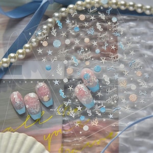 May include: Clear sheet of nail art stickers with a seaside theme. Designs include seahorses, seashells, starfish, and bows in pastel blue, peach, and white. Some nails are painted with a gradient of pink and blue, with the words "Pelagic" and "Seaside".