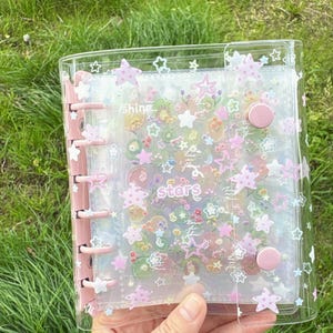 May include: Clear, plastic binder with a pink spine and closures. The binder is decorated with star-shaped stickers and the word "Stars". The inside pages are filled with colorful floral and star-shaped stickers. The binder is held in front of a green background.