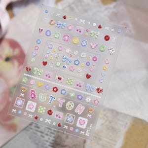 May include: A translucent sticker sheet featuring a variety of small decorative icons including colorful buttons, bows, hearts, flowers, strawberries, and teddy bear faces. The word BUTTON is printed in large, patterned letters across the center.