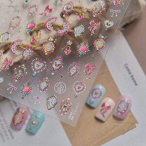 May include: A transparent sheet of adhesive nail art stickers featuring gold-rimmed designs of clocks, pink bows, hearts, and ornate frames. The sheet includes small star accents and text reading 5805. Several stickers are applied to painted nail tips.