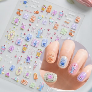 Puede incluir: Una hoja de pegatinas adhesivas para uñas con varios diseños de temática veraniega que incluyen helados, frutas y bebidas, con texto que dice Summer is calling, orange, peach y blueberry, junto a uñas decoradas con estas pegatinas.