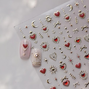 Peut inclure: Feuille d'autocollants dorés pour nail art avec des cœurs, des soleils, des lunes, des étoiles et des nœuds avec des accents rouges. Deux ongles sont décorés de motifs similaires. Les autocollants sont sur un support transparent.