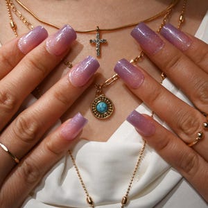 May include: Close-up of a set of long, square-shaped artificial nails with a gradient design, transitioning from a light pink at the base to a lavender purple at the tips. The nails are adorned with a subtle glitter effect. Gold necklaces and rings are also visible.