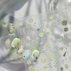 May include: A sheet of adhesive nail decals featuring various green apple motifs, four-leaf clovers, bows, stars, music notes, and beverage glasses. Several individual decals are applied to artificial nail tips, showcasing the designs.