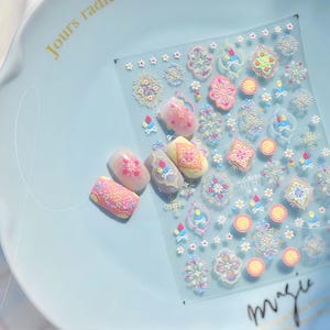 Op de afbeelding: Een vel met nagelkunststickers met diverse kleurrijke bloemen- en geometrische ontwerpen. Er liggen meerdere versierde nagels in de buurt van het vel. De ontwerpen bevatten roze, gele, blauwe en groene elementen. Het vel is transparant en ligt op een lichtblauwe ondergrond.