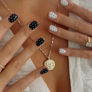 Puede incluir: Primer plano de manos con manicura de lunares en blanco y negro. También se ven un collar de oro con un colgante circular y un collar de conchas. Un anillo de oro con una piedra grande y transparente está en un dedo.