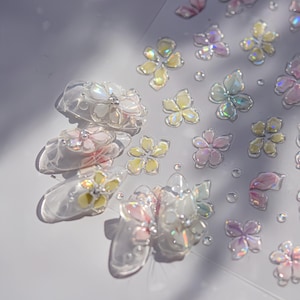 May include: A selection of iridescent flower nail art decorations in shades of yellow, pink, and white. The nail art is displayed on a clear sheet, with some pieces appearing on a nail. The flowers have a three-dimensional appearance.