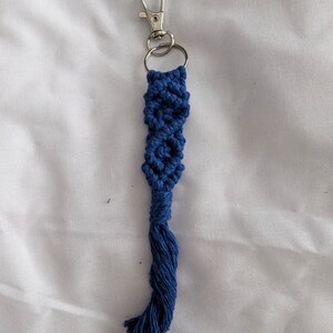 Puede incluir: Un llavero de macramé hecho a mano en color azul oscuro. El llavero presenta un diseño tejido con un cierre y un anillo de color plateado. La parte inferior del llavero tiene una borla de hilos sueltos.