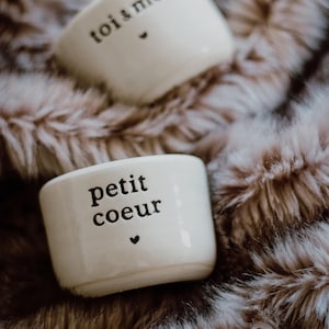 Peut inclure: Deux petits gobelets en céramique blanc cassé avec du texte noir et un symbole de cœur. L'un des gobelets porte l'inscription "toi & moi" et l'autre "petit coeur". Les gobelets reposent sur une surface en fausse fourrure marron.