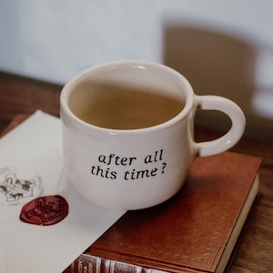 Può includere: Una tazza in ceramica color crema con un manico arrotondato e il testo nero "after all this time?" stampato sul davanti. La tazza è posizionata sopra un libro rilegato in pelle marrone accanto a una busta bianca con un sigillo in ceralacca rossa.