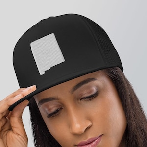 May include: Black trucker hat featuring a white embroidered outline of the state of New Mexico. The hat has a flat brim and a mesh back. The hat is being worn by a person.